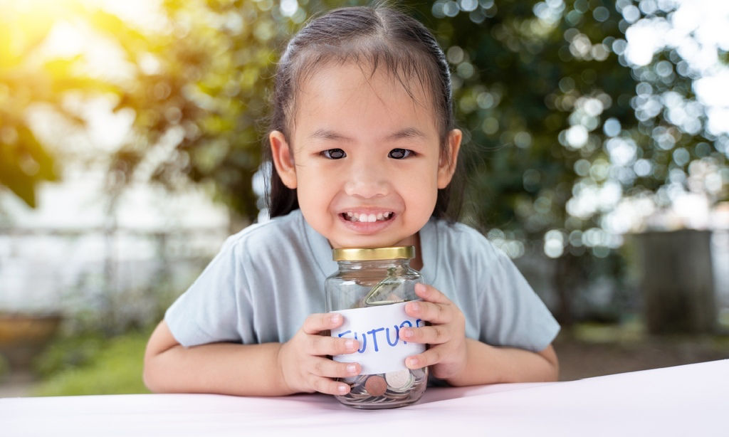 A young Asian girl places her chin on top of a coin-filled jar with the label future.