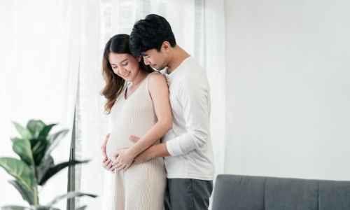 Young Asian husband hugs wife from behind and cradles her pregnant belly.