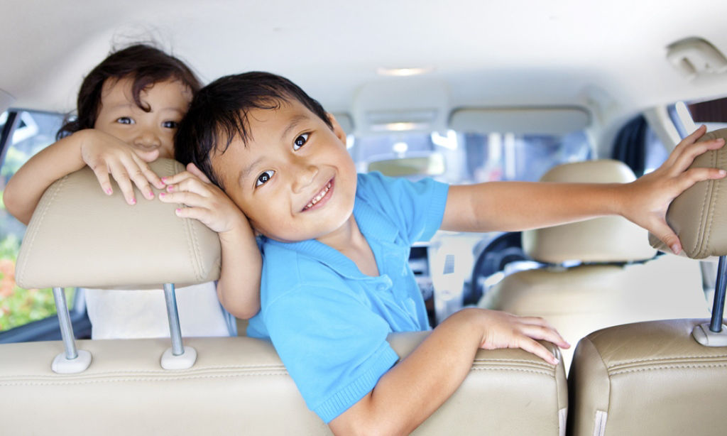 Two preschool-age Asian kids pose for the camera while at a car back seat.