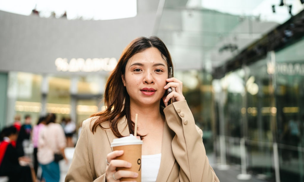 Young Asian professional talks on her smartphone while holding a cup of coffee.