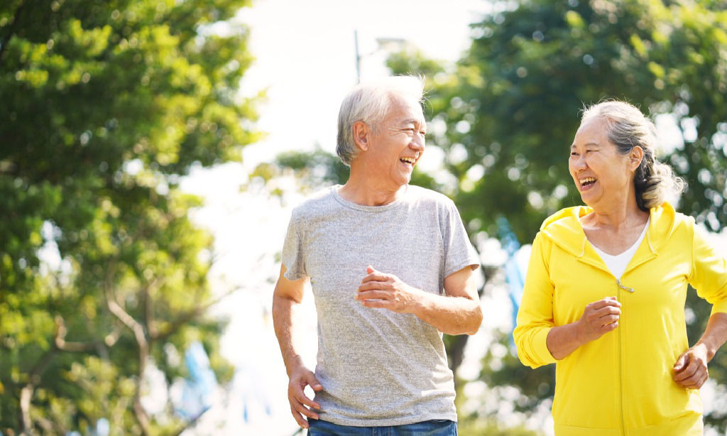 An Asian couple in their 60s smile at each other as they jog outdoors.