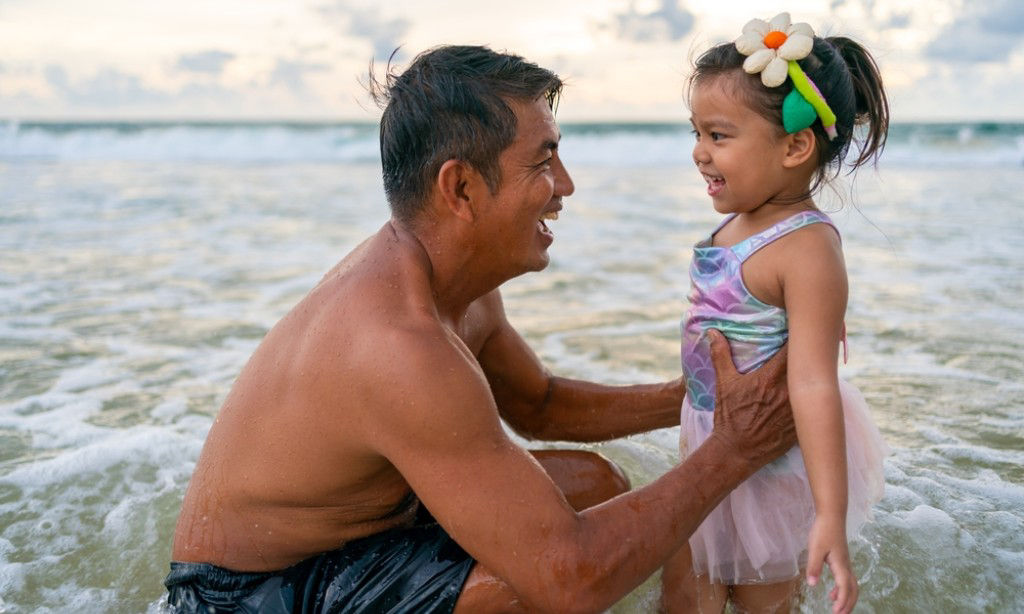 Laughing Asian dad gets down to his daughter’s eye-level at the beach.