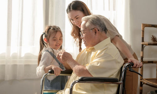 Mother and daughter show an origami bird to a grandfather sitting in a wheelchair